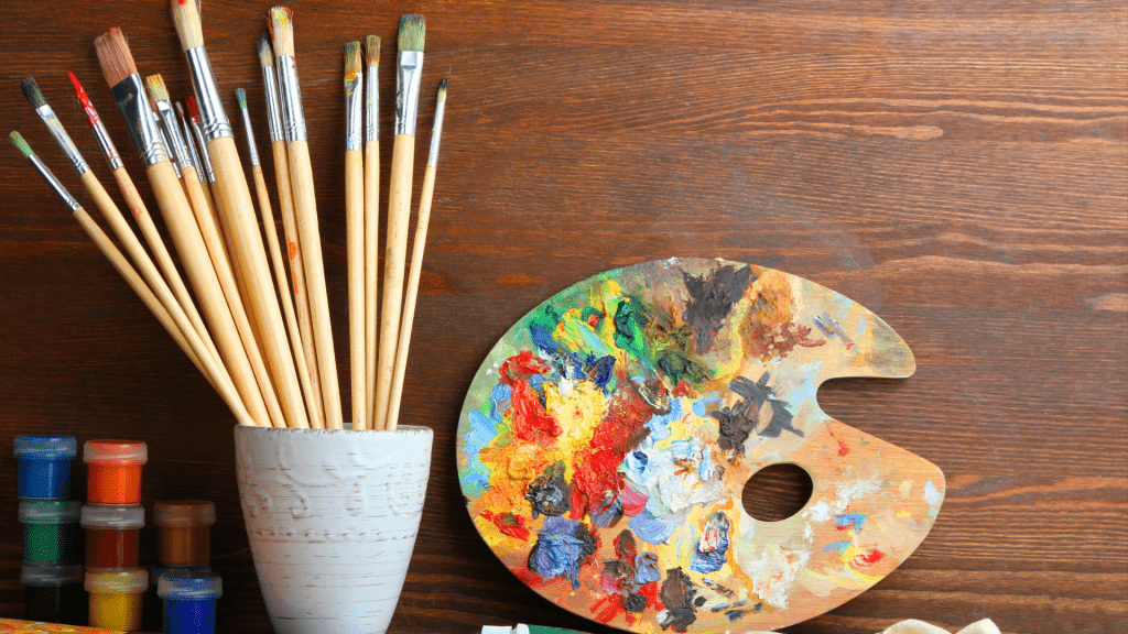 A wooden palette covered in colorful paint alongside various paintbrushes, highlighting the traditional artistic process in "Art vs. Design in Creative Industry.